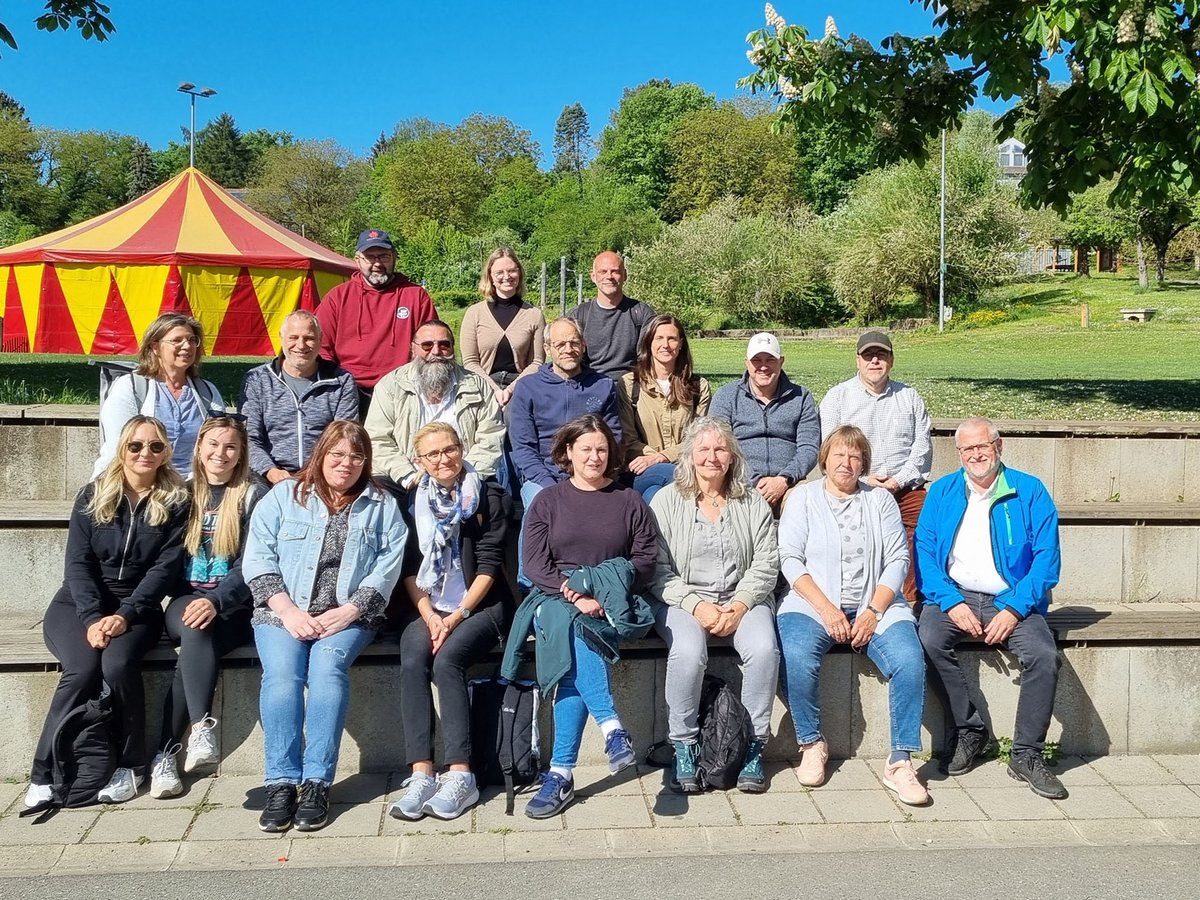 Eine Gruppe Erwachsener sitzt auf Stufen im Freien vor einem rot-gelben Zirkuszelt und Bäumen.