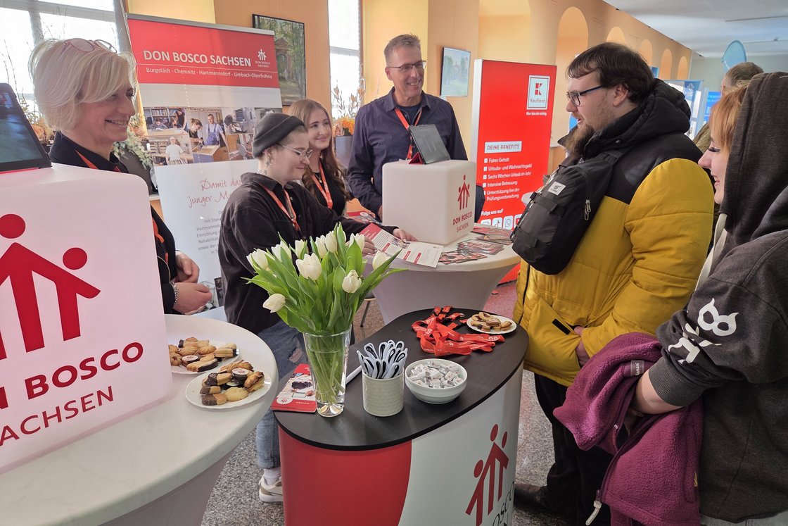 Mehrere Personen unterhalten sich an Messeständen von Don Bosco Sachsen und Kaufland, umgeben von Infomaterial und Snacks.