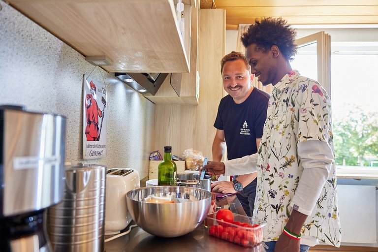 Zwei Männer kochen zusammen in einer Küche, einer lächelt, der andere rührt in einem Topf. Es gibt Küchengeräte und Zutaten.