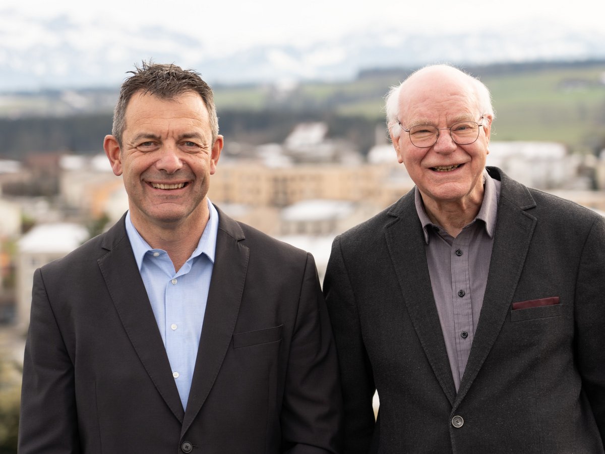 Zwei lächelnde Männer, ein jüngerer und ein älterer, stehen nebeneinander im Freien. Im unscharfen Hintergrund sind eine Stadt und Berge zu sehen.