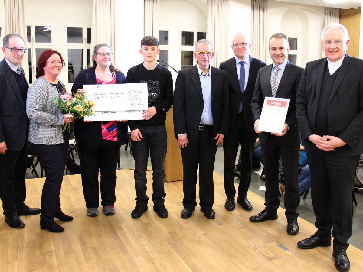 Neun Personen, darunter zwei Frauen mit Blumen und Scheck sowie sieben Männer mit einer Urkunde, bei einer feierlichen Übergabe.