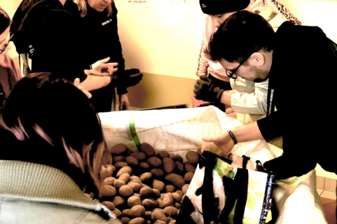 Menschen versammeln sich um einen großen Kartoffelsack, eine Person füllt eine Einkaufstasche, vor Wandskizzen.