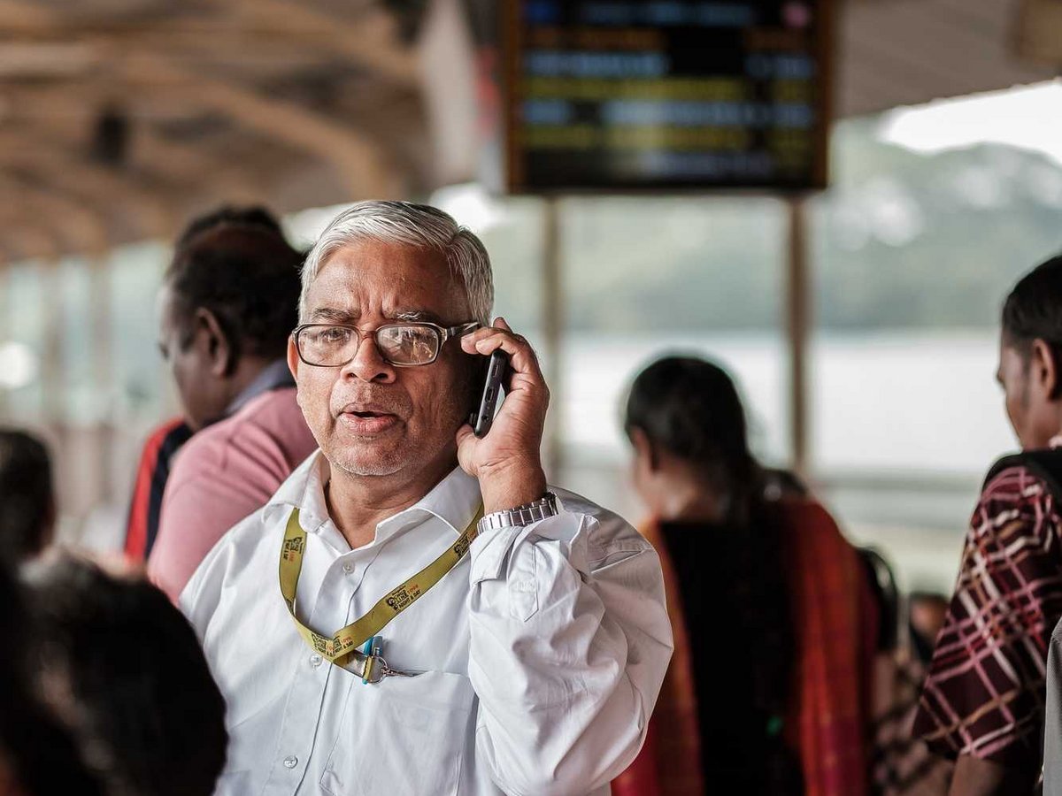 Ein älterer Mann mit Brille telefoniert auf einem belebten Bahnhofsgleis, über ihm ein Schild mit „PLATFORM 4“.