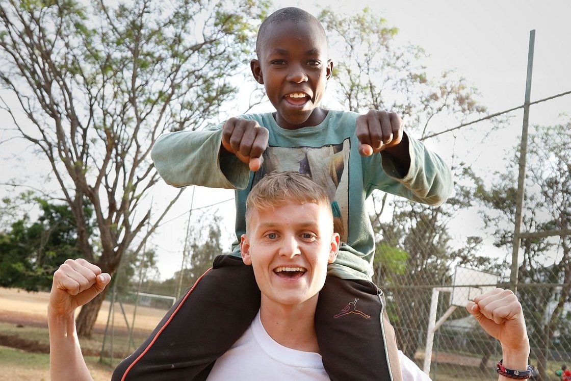 Ein lächelnder schwarzer Junge sitzt auf den Schultern eines fröhlichen weißen Mannes, beide heben spielerisch die Fäuste auf einem Sportplatz im Freien.