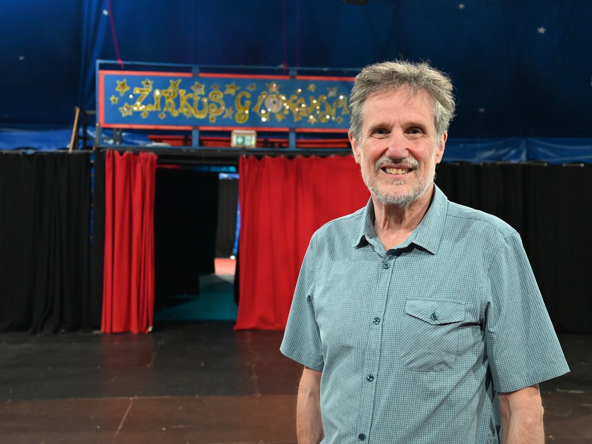 Lächelnder Mann im blau karierten Hemd in einem Zirkuszelt mit Zirkusschild und roten Vorhängen im Hintergrund.