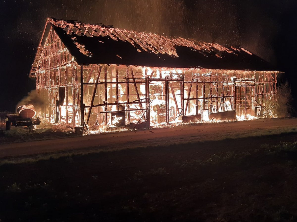 Eine große Holzscheune, die nachts vollständig in intensiven Flammen steht, das Dach ist abgebrannt und Funken steigen auf.