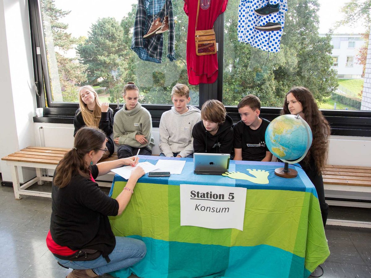 Schüler und eine Frau an der 'Station 5 Konsum' mit Globus, Laptop und an der Fensterscheibe hängender Kleidung.