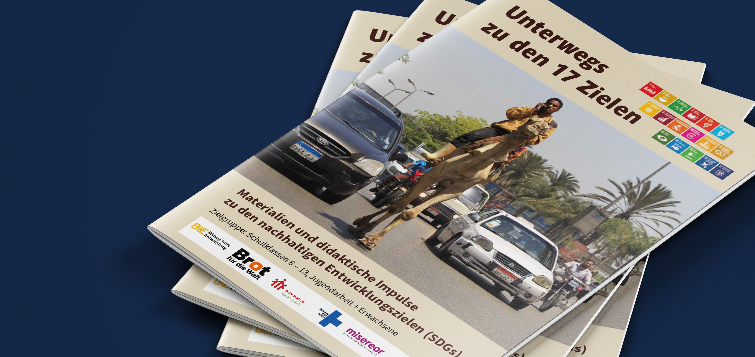 Stapel "Unterwegs zu den 17 Zielen"-Broschüren; Titelbild: Mann auf Kamel im Straßenverkehr mit Autos und Palmen.