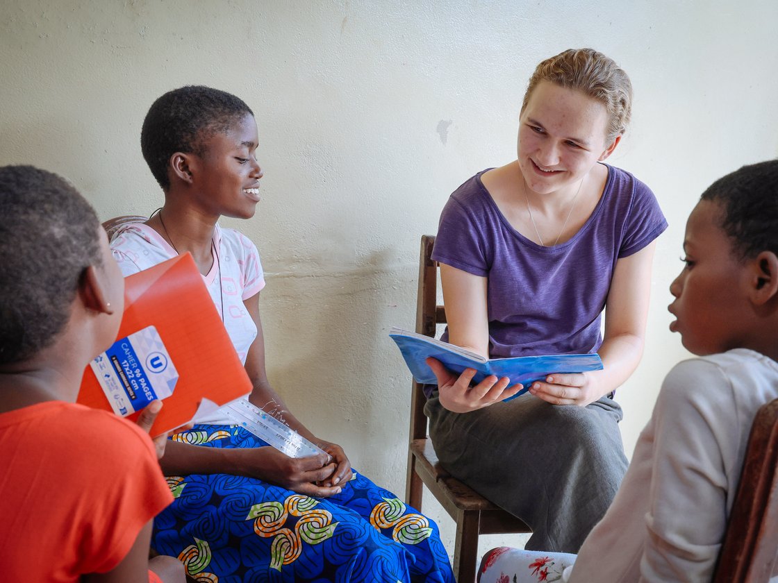 Eine lächelnde Frau liest zwei Kindern ein Buch vor, während eine andere Frau eine Mappe hält. Alle sind in eine Lernstunde vertieft.