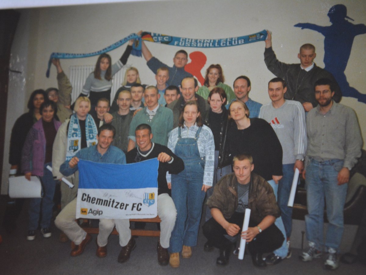 Eine Gruppe von Chemnitzer FC-Fans hält Schals und ein Banner und posiert für ein Foto im Innenbereich mit Vereinsdekor.