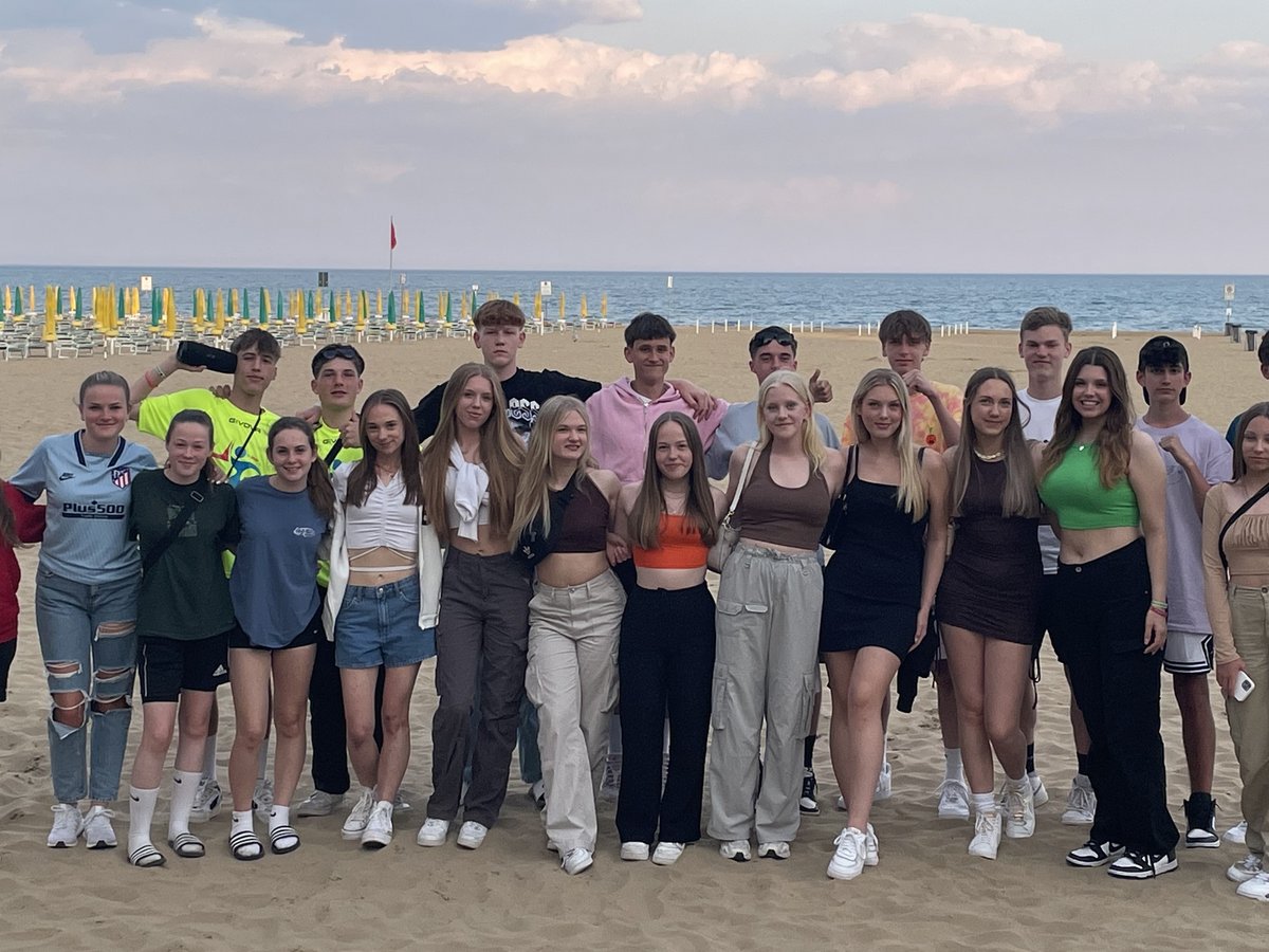 Eine große Gruppe lächelnder junger Leute, hauptsächlich Teenager, steht am Sandstrand mit Meer und bunten Sonnenschirmen.