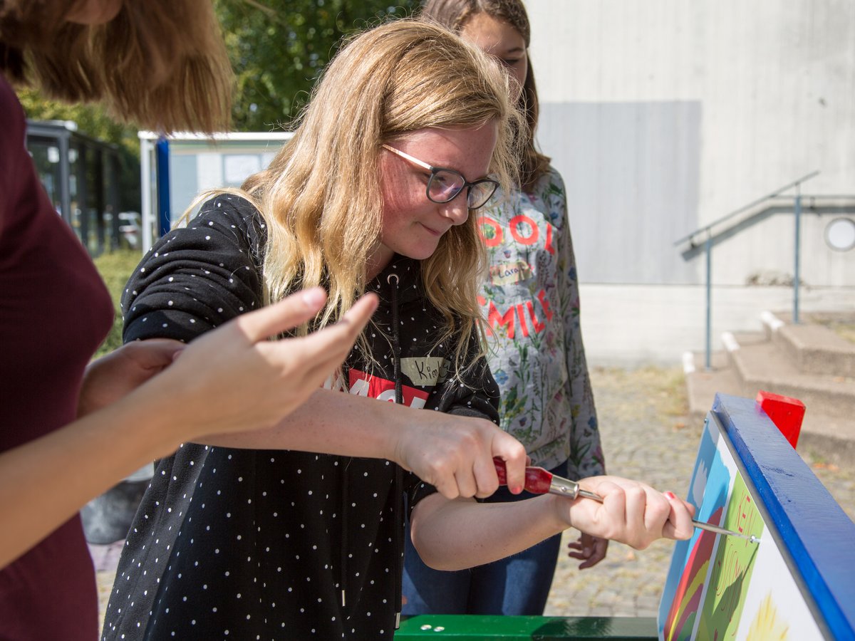 Junge Frau mit Brille schraubt im Freien an einem farbenfrohen Brett. Hände einer anderen Person sind im Vordergrund sichtbar.