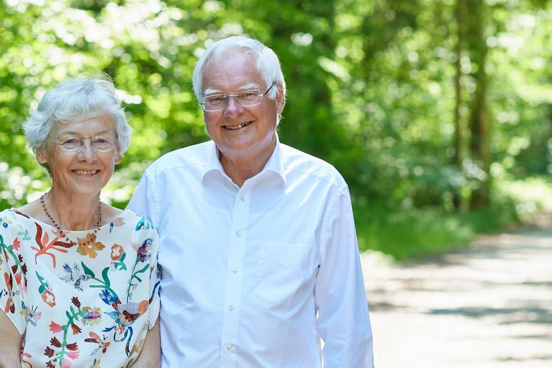 Ein älteres Ehepaar lächelt in einem sonnigen Park mit grünen Bäumen und einem Weg.