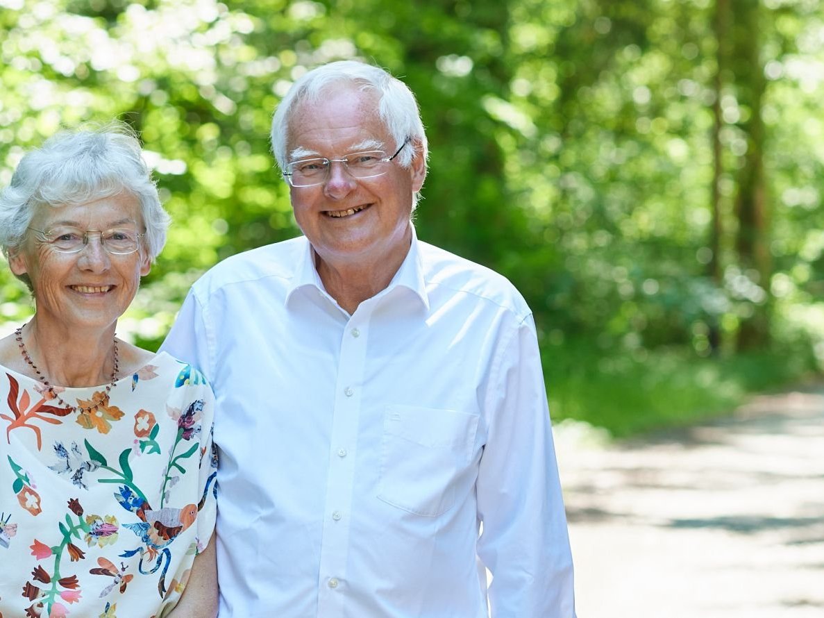Ein älteres Ehepaar lächelt in einem sonnigen Park mit grünen Bäumen und einem Weg.