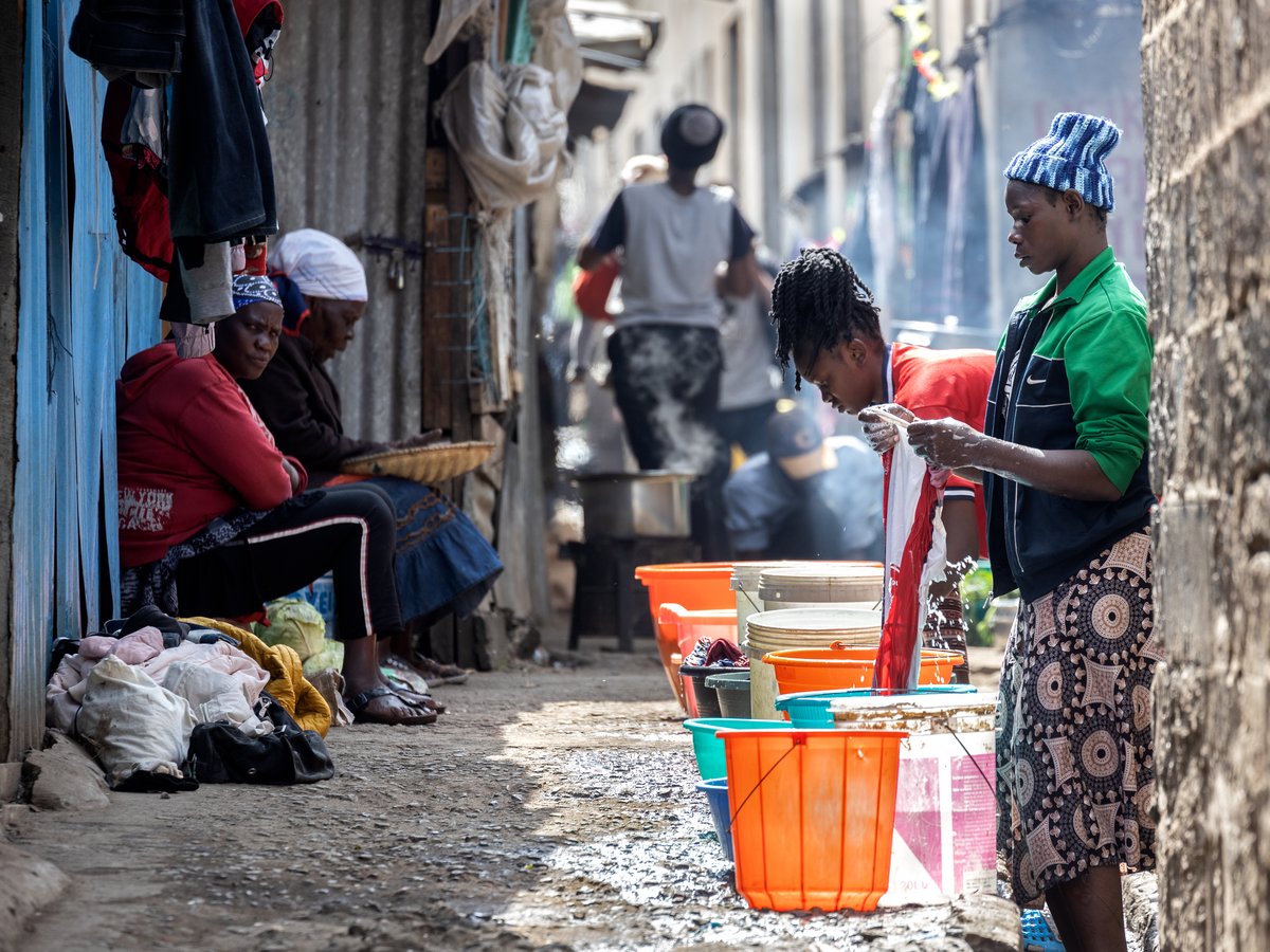 Frauen waschen Kleidung in bunten Eimern und sitzen an einem Weg in einer lebhaften, sonnigen Gemeinschaft mit Wellblechwänden.