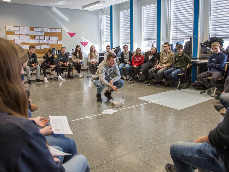 Schüler in einem Klassenraum-Workshop, ein junger Mann demonstriert eine Aktivität in der Mitte, umgeben von sitzenden Mitschülern.