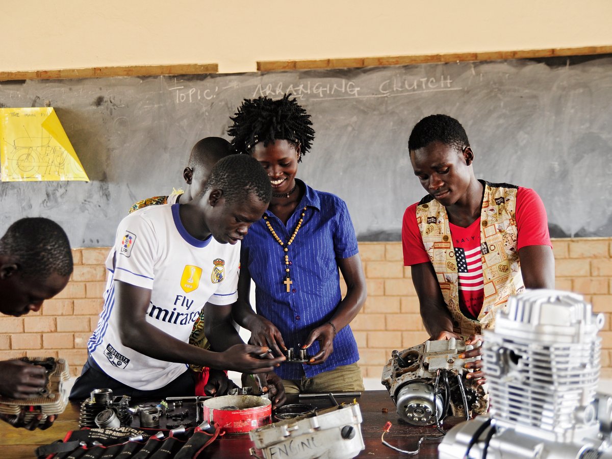 Schüler in einem afrikanischen Berufskurs üben Motorradmechanik, montieren Teile auf einem Tisch unter einer Tafel mit 'ARRANGING CLUTCH'.