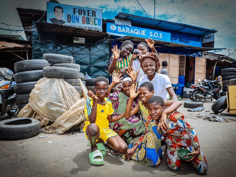 Lachende afrikanische Kinder und Jugendliche winken vor 'FOYER DON BOSCO' und 'BARAQUE S.O.S' Gebäuden, umgeben von Altreifen.
