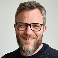 Ein lächelnder Mann mit Bart und Brille in einem dunklen Pullover blickt direkt in die Kamera.