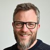 Ein lächelnder Mann mit Bart und Brille in einem dunklen Pullover blickt direkt in die Kamera.