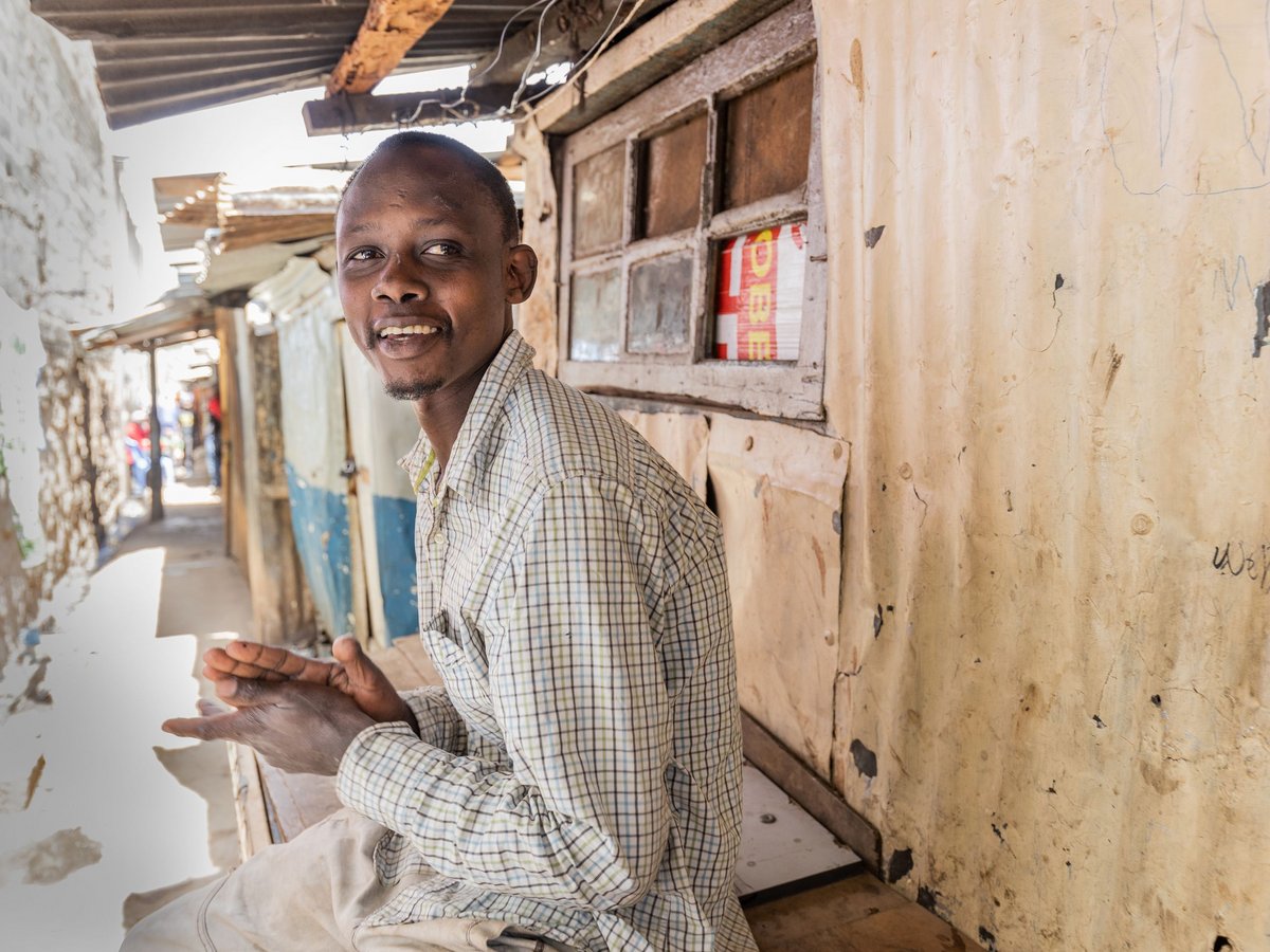 Ein lächelnder afrikanischer Mann in einem karierten Hemd sitzt auf einer Bank in einer sonnigen Gasse und blickt in die Kamera.