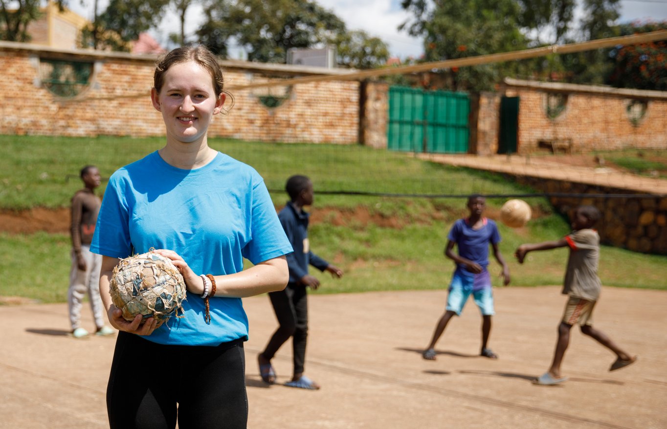 Eine junge Frau im blauen Shirt lächelt und hält einen improvisierten Ball, während Kinder im Hintergrund auf einem Sandplatz spielen.