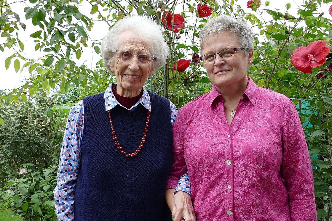 Eine ältere Frau und eine mittelalte Frau lächeln und stehen Arm in Arm in einem Garten mit roten und rosa Blumen.