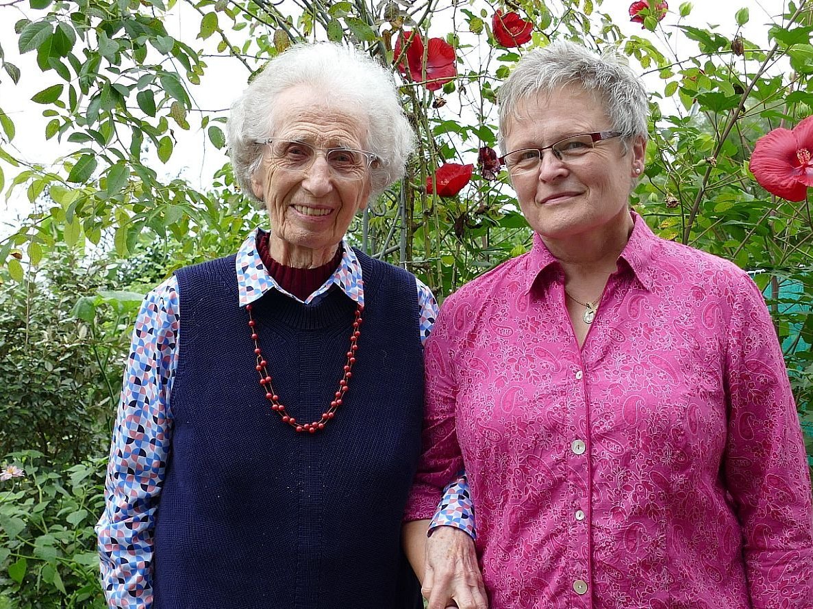 Eine ältere Frau und eine mittelalte Frau lächeln und stehen Arm in Arm in einem Garten mit roten und rosa Blumen.