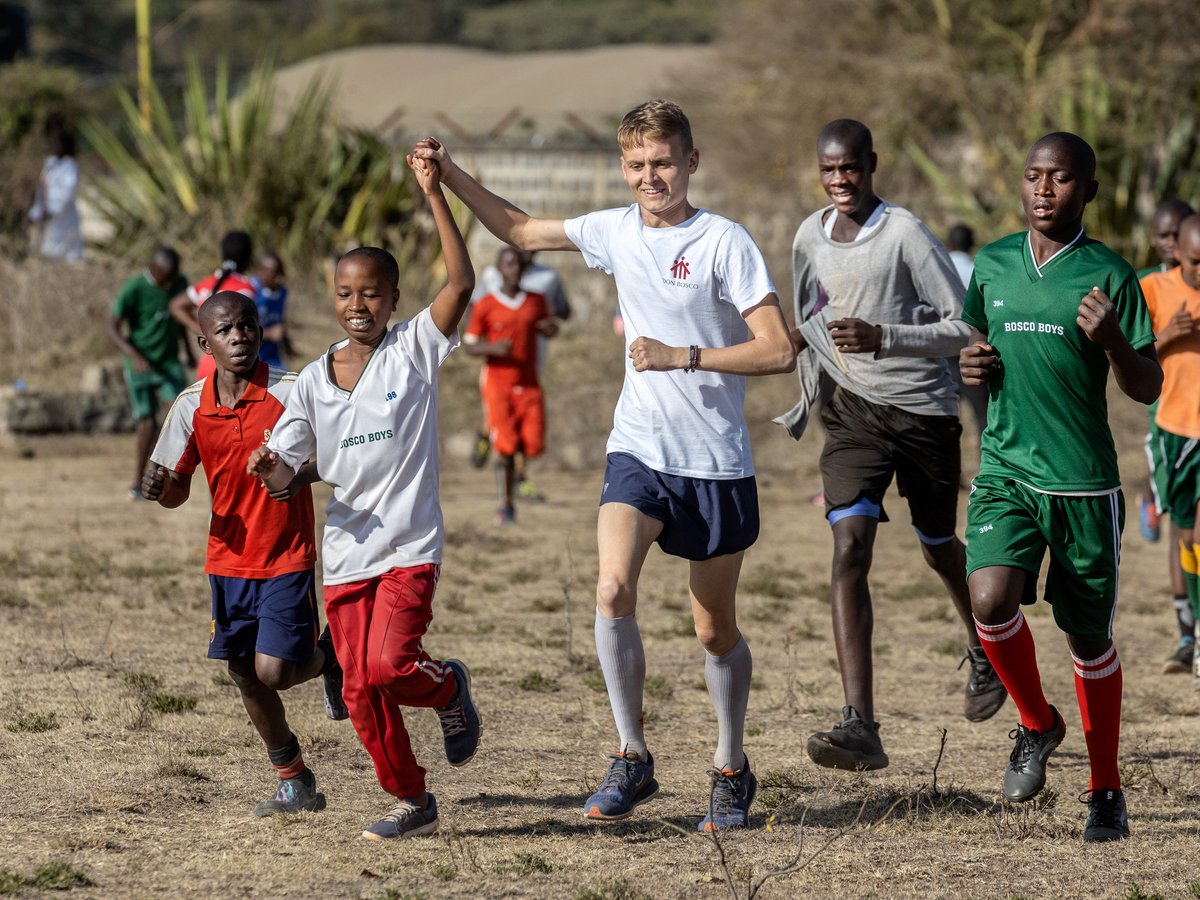 Diverse Jungen rennen freudig auf trockenem Feld. Zwei Jungen in der Mitte halten Hände und lächeln.