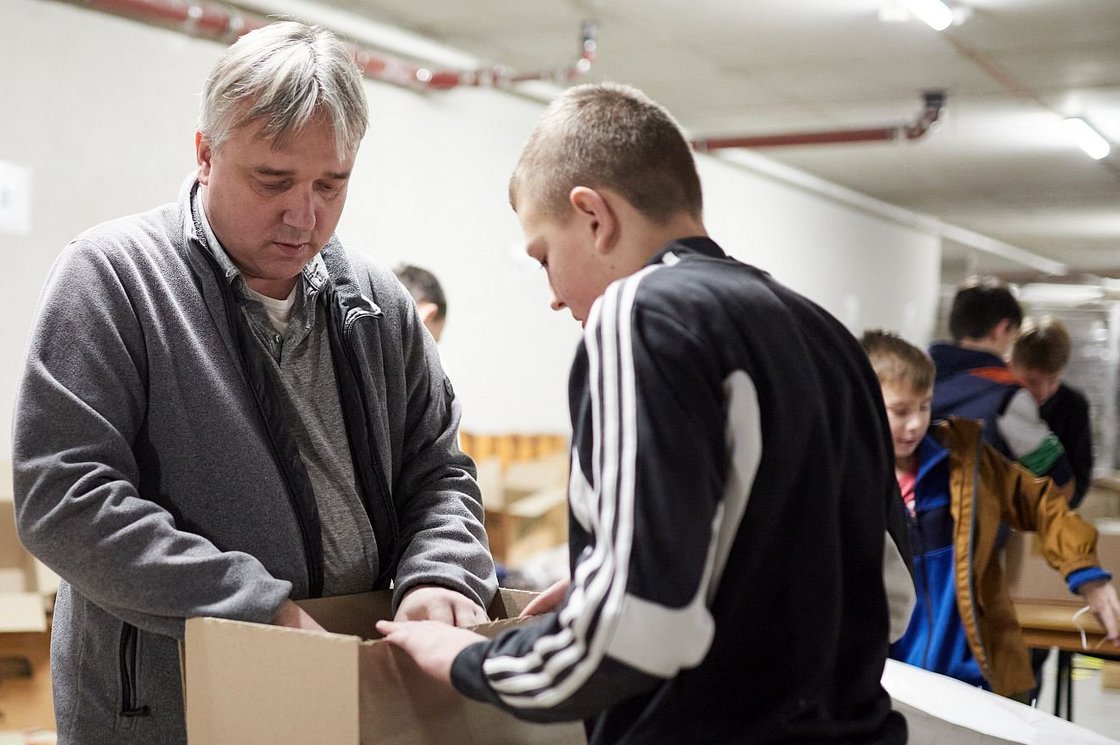 Mann und Junge packen Hilfsgüter in Kartons in einem Lagerraum, weitere Kinder helfen im Hintergrund.