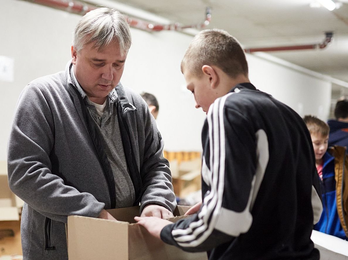Mann und Junge packen Hilfsgüter in Kartons in einem Lagerraum, weitere Kinder helfen im Hintergrund.