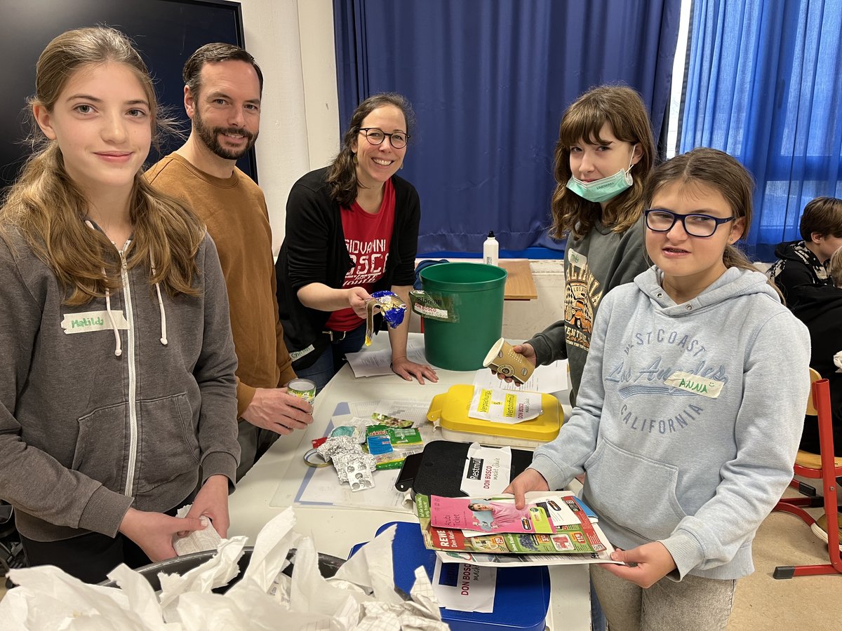 Erwachsene und Kinder lernen beim Abfallsortieren: Eine Gruppe lächelt beim Recycling von Papier, Plastik und Metall in die Kamera.