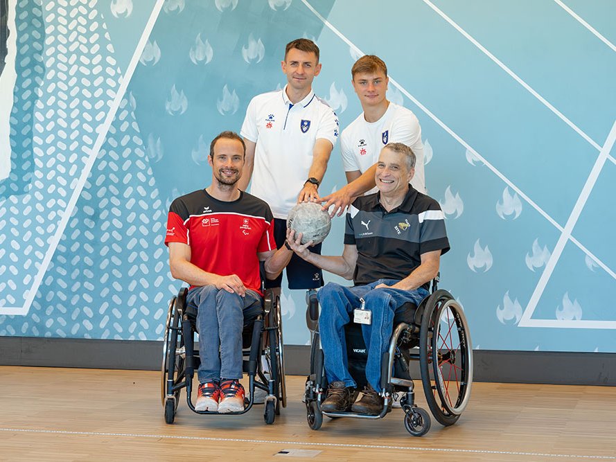 Vier Männer, zwei im Rollstuhl und zwei stehend, lächeln und halten gemeinsam einen grauen Sportball. Symbol der Einheit im Sport.
