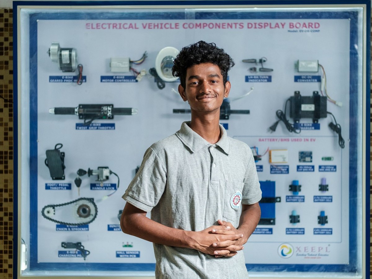 Lächelnder junger Mann in grauem Poloshirt vor einer Anzeigetafel für Elektrofahrzeugkomponenten.