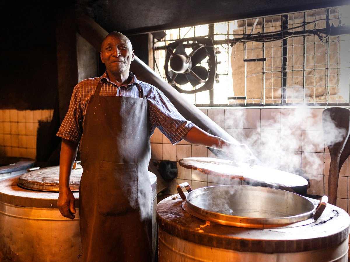 Ein lächelnder Mann in einer Küche hebt den Deckel eines dampfenden Topfes an. Um ihn herum stehen große Behälter.