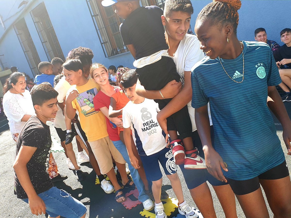 Fröhliche, vielfältige Gruppe von Kindern und Erwachsenen genießt ein Outdoor-Spiel, stehend in einer Reihe auf bunten Puzzlematten.