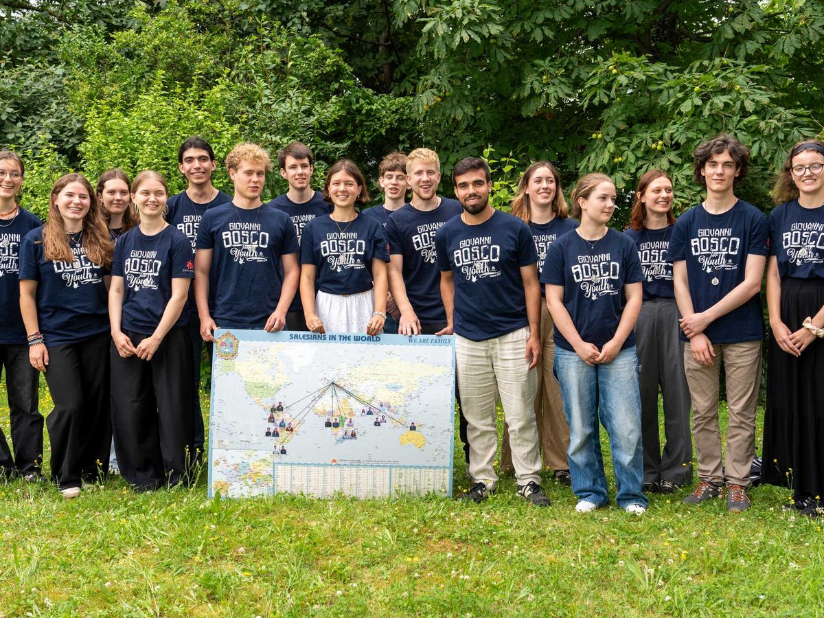 Vielfältige Jugendliche in 'Giovanni Bosco Youth'-T-Shirts posieren mit einer Weltkarte 'Salesianer in der Welt' auf einer Wiese.