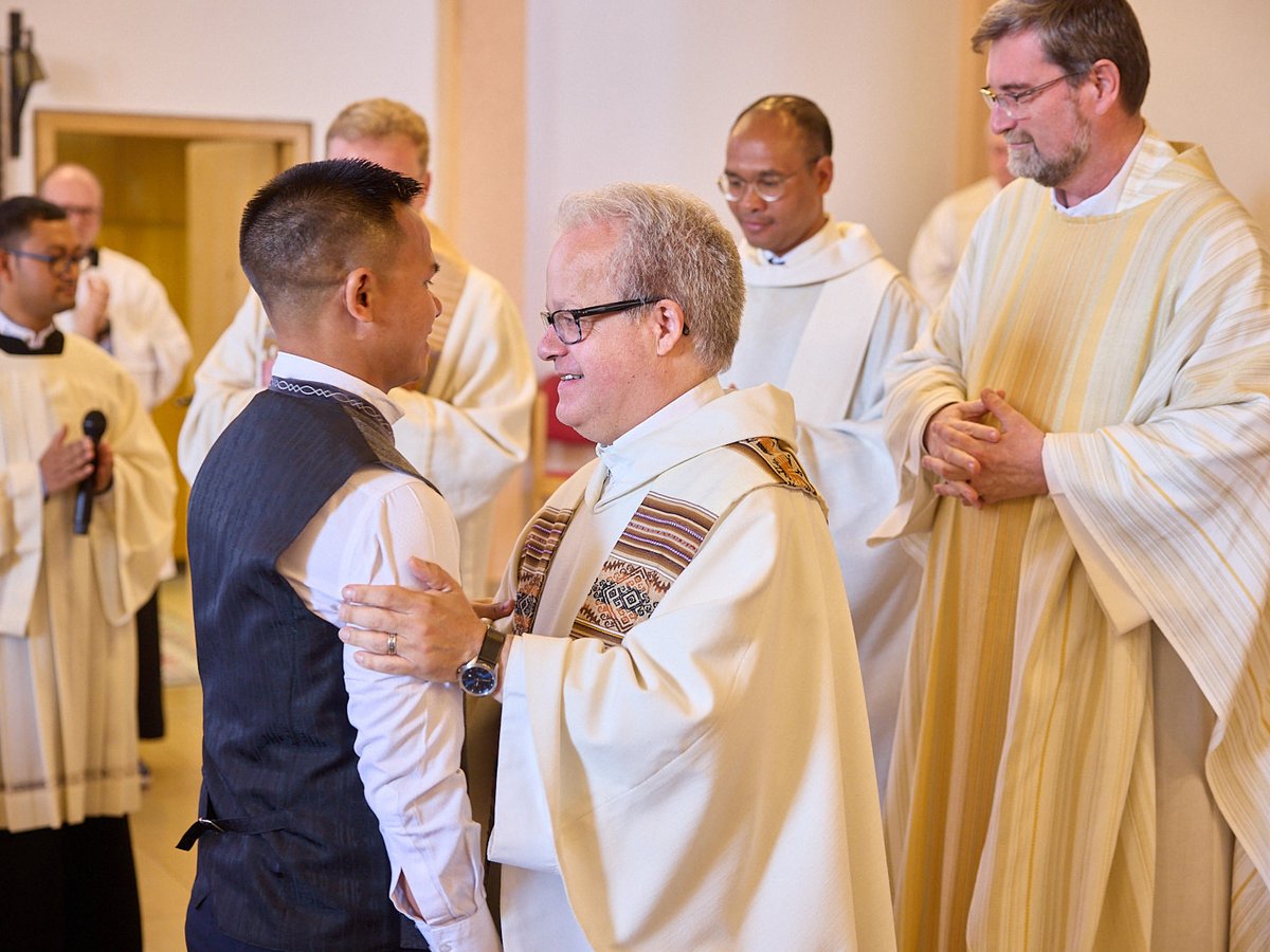 Geistliche bei einer Kirchenzeremonie Priester in weißen Gewändern legt Hand auf Arm eines lächelnden Mannes in Weste während einer Zeremonie; andere Geistliche beobachten.