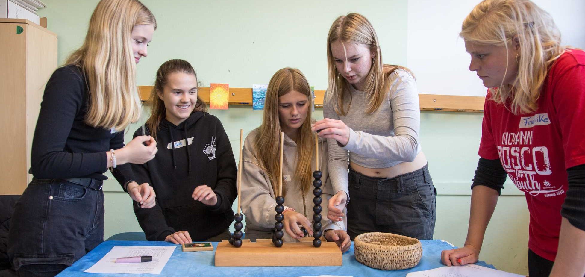 Fünf junge Frauen an einem Tisch mit blauem Tuch, die an einer Station zum CO2-Ausstoß-Vergleich mit einem Abakus arbeiten.
