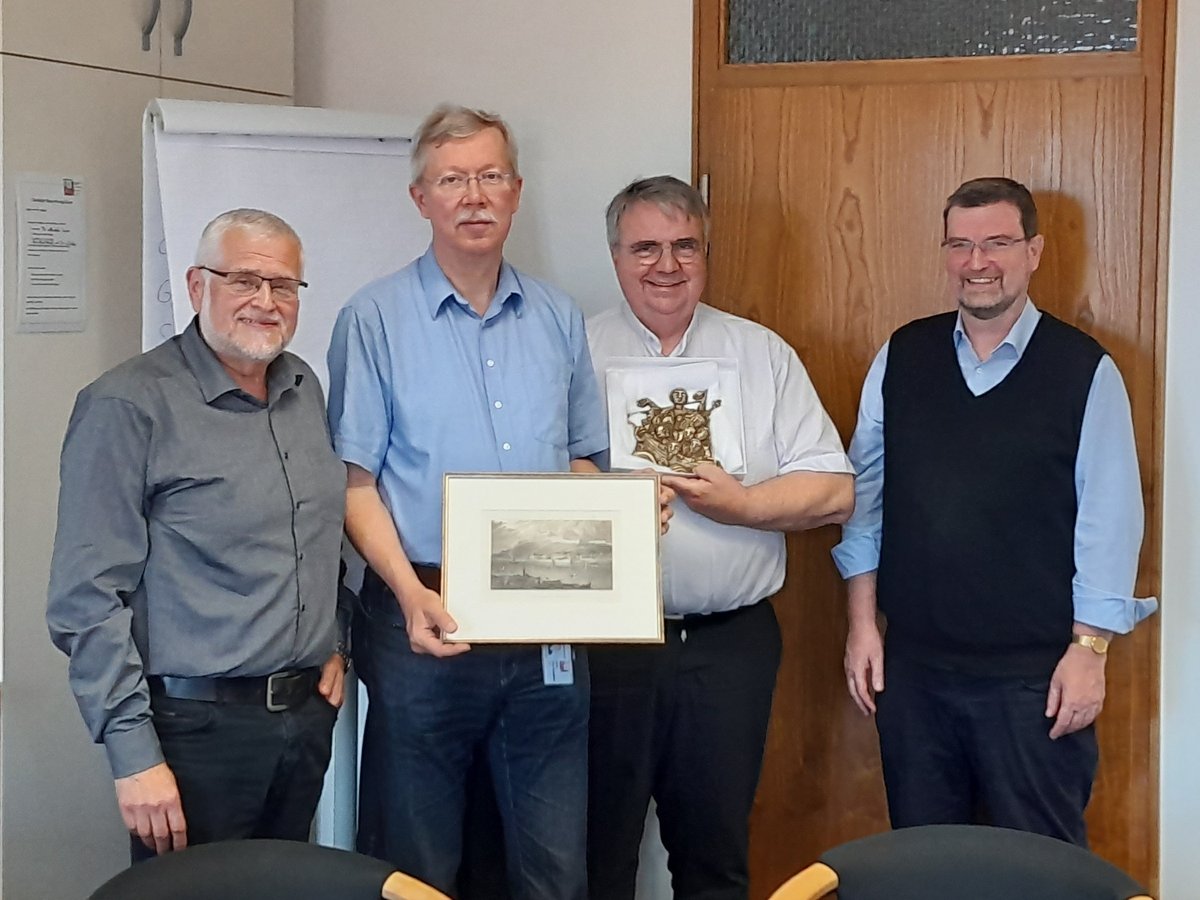 Vier lächelnde Männer im Büro, zwei halten gerahmte Objekte: ein Seestück und ein goldenes Relief.