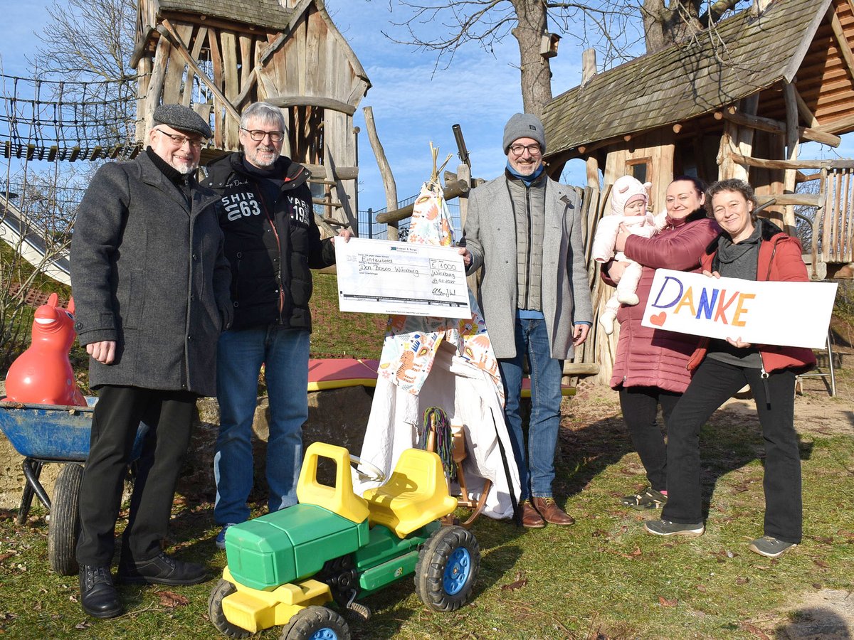 Gruppe von Erwachsenen und Baby auf Spielplatz; Männer halten Spendenscheck, Frauen DANKESchild, im Hintergrund Holzspielgeräte.