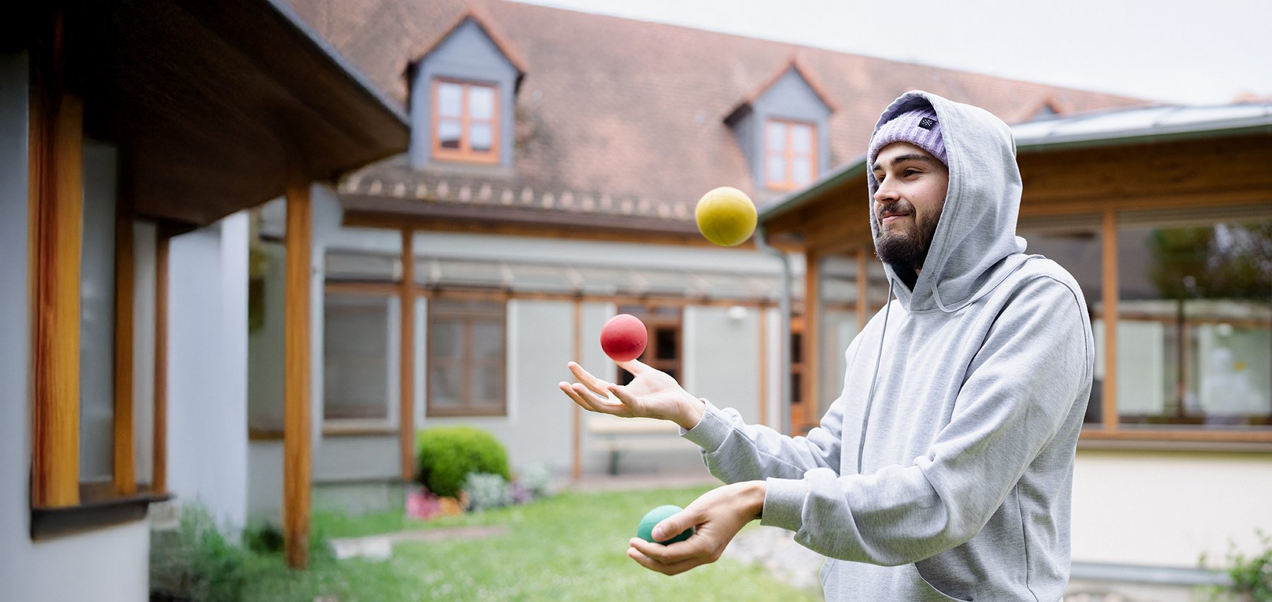 Ein bärtiger Mann in grauem Hoodie und lila Mütze jongliert drei bunte Bälle (gelb, rot, grün) in einem Hof.