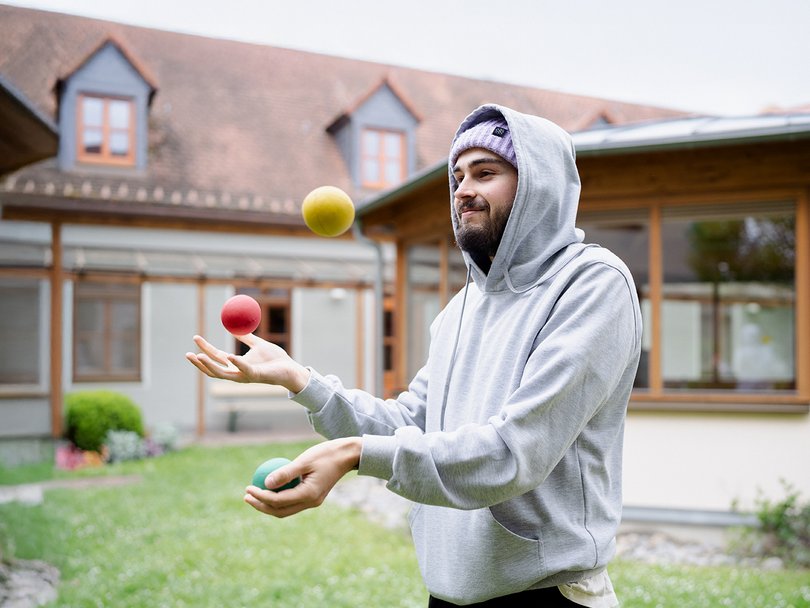 Ein bärtiger Mann in grauem Hoodie und lila Mütze jongliert drei bunte Bälle (gelb, rot, grün) in einem Hof.