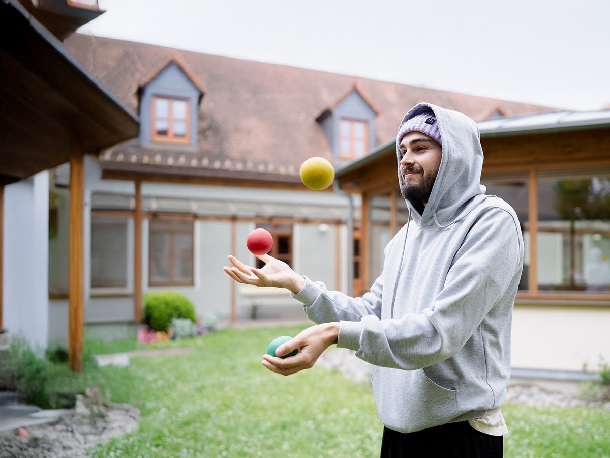 Ein bärtiger Mann in grauem Hoodie und lila Mütze jongliert drei bunte Bälle (gelb, rot, grün) in einem Hof.