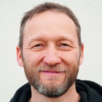 Nahaufnahme eines lächelnden Mannes mit braun-grauem Bart und Haaren, der einen schwarzen Kapuzenpullover trägt.
