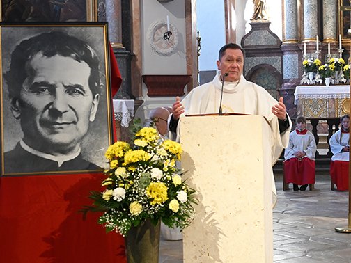 Ein Priester predigt in einer Kirche, neben einem Porträt von Don Bosco und einem Blumenarrangement.