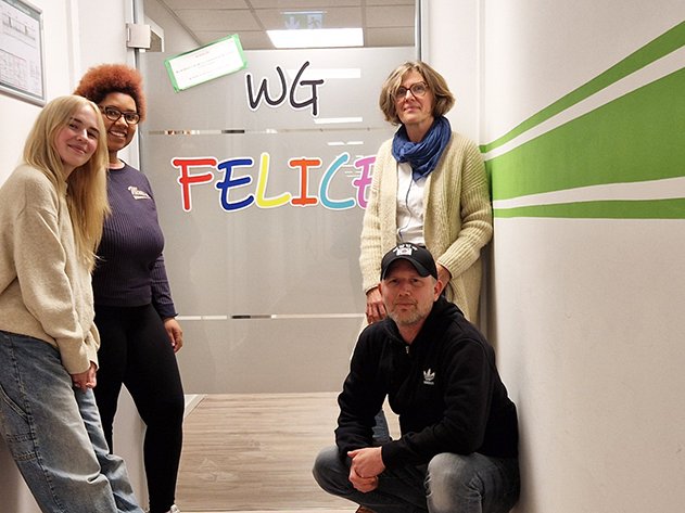 Vier Personen (3 Frauen, 1 Mann) posieren in einem Flur. Eine Glastür mit 'WG FELICE' ist hinter ihnen, und eine Wand mit grünen Streifen.