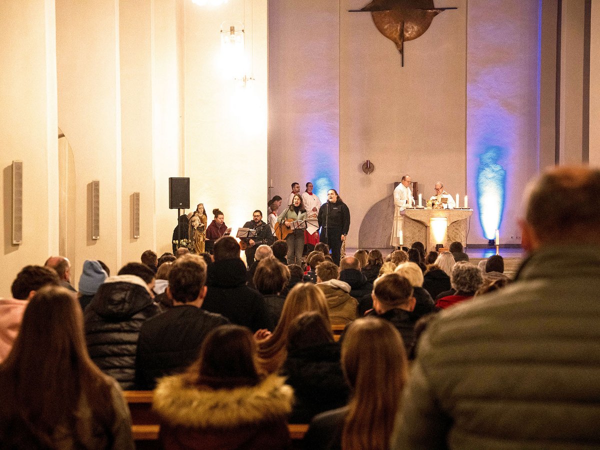 Eine Band spielt Musik in einer beleuchteten Kirche, ein Priester steht am Altar, während das Publikum lauscht.