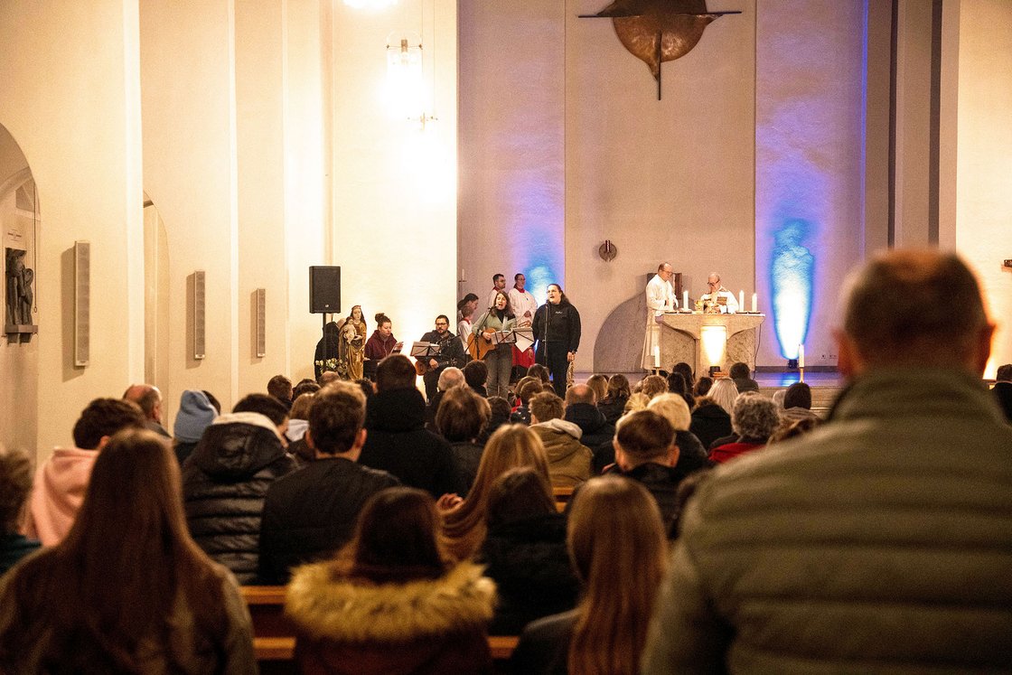 Eine Band spielt Musik in einer beleuchteten Kirche, ein Priester steht am Altar, während das Publikum lauscht.