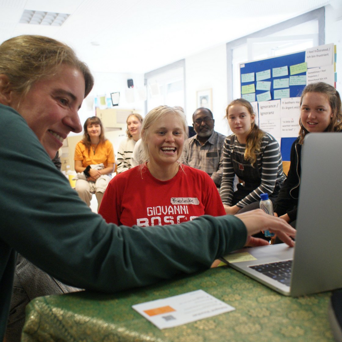 Eine vielfältige Gruppe lächelt und blickt auf eine Person, die einen Laptop in einem Klassenzimmer oder Workshop nutzt.